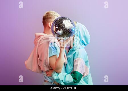 Blonde Mann in rosa Hoodie und Frau in stilvollen Mint Bomber küssen gekleidet, Gesichter unbesieht hinter funkelnden Disco Ball, posiert im Studio über pu Stockfoto