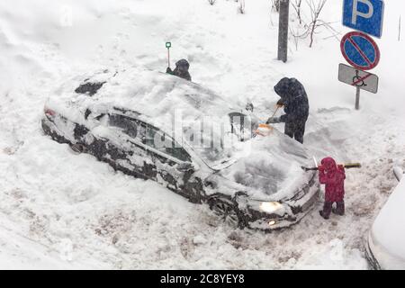 Nicht erkennbare Menschen räumen schneebedeckten Auto in Schneesturm. Kinder helfen ihrem Vater, Schnee vom Auto zu räumen. Es gibt starken Schneesturm outsid Stockfoto