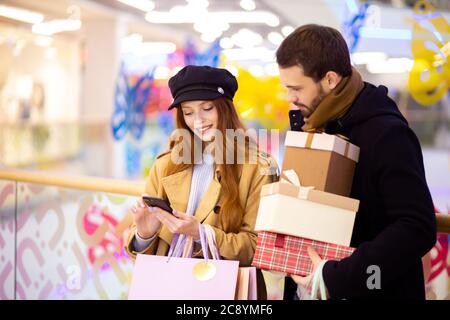 Rothaarige kaukasische Frau mit Einkaufstaschen zeigen etwas im Smartphone bärtigen Mann hält Geschenke, diskutieren Einkaufen innerhalb der Mall Stockfoto