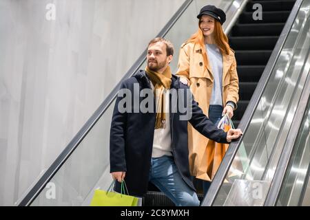 Glückliches junges kaukasisches Paar mit Einkaufstaschen, die auf der Rolltreppe aufsteigen und sich das Einkaufszentrum, Shopaholics, angucken Stockfoto