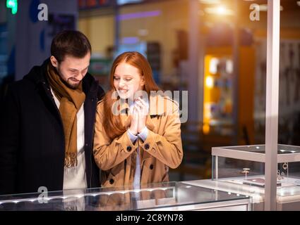 Frisch verheiratetes Paar, kaukasischer Mann und Frau beim Einkaufen in der Mall, wählen Sie teuren Schmuck für Frauen, tragen Mantel und umarmenden Mann Stockfoto