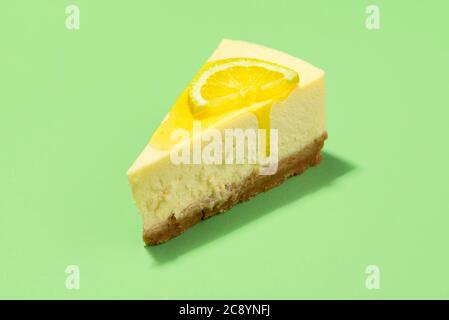 Käsekuchen-Scheibe mit Zitronensauce tropfend und Zitronenscheibe, isoliert auf grünem Hintergrund. Nahaufnahme einer Scheibe Käsekuchen mit gelbem Sirup Belag. Stockfoto