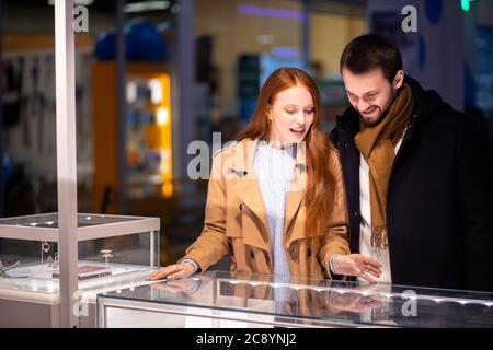 Schönes Paar, das im Geschäft teuren goldenen oder silbernen Schmuck auswählt, lächelnde glückliche Frau mit roten Haaren und bärtiger Mann, der Mantel trägt Stockfoto