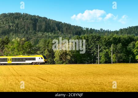 Elektrozug durch landwirtschaftliche Felder und Wald, bei Sonnenaufgang, in Schwabisch Hall, Deutschland. Umweltfreundlicher gelber Personenzug. Stockfoto