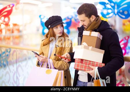 Rothaarige kaukasische Frau mit Einkaufstaschen zeigen etwas im Smartphone bärtigen Mann hält Geschenke, diskutieren Einkaufen innerhalb der Mall Stockfoto