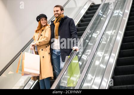 kaukasische rothaarige Frau und bärtiger Mann lächeln, genießen Sie den Einkauf zusammen, happ nach dem Kauf neuer Kleidung und Dinge. Steigt auf Rolltreppe Stockfoto