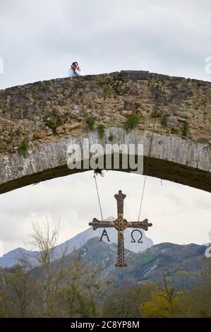 Fotograf über der Steinbrücke, die fälschlicherweise als römische Brücke bezeichnet wird, an der das Kreuz Cruz de la Victoria hängt (Cangas de Onís, Asturias, Spanien) Stockfoto