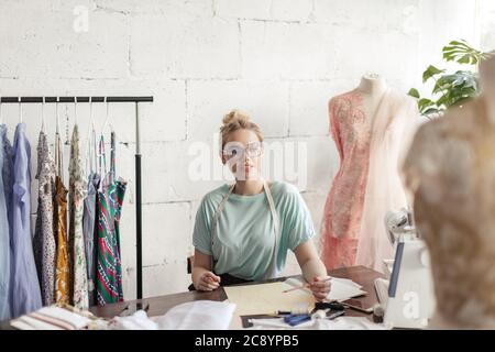 Kreative weibliche Näherin in Brillen und Bandlaufwerke arbeiten am Design der neuen Kleid Skizzen am Desktop sitzen in Vintage stilvolle Atelier Stockfoto