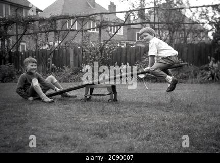 1950, historisch, zwei junge Jungen spielen in einem Garten auf einer hausgemachten Wippe, ein Brett aus Holz auf der Basis eines alten Metall vierrädrigen Spielzeug gelegt. In Parks und Spielplätzen üblich, machen Wippen Spaß, helfen beim Muskelaufbau und fördern das kooperative Spielen zwischen zwei Kindern. Was für ein Spaß mit Phantasie und einem schmalen Holzspank! Stockfoto