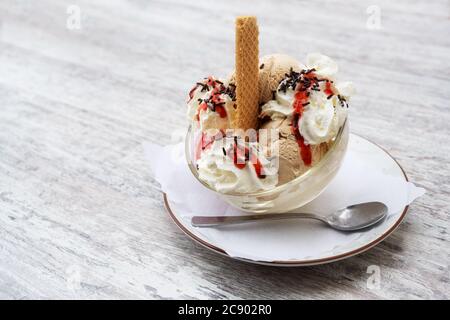Eisbecher mit Eisschaufeln, Schlagsahne, rote Obstsoße und Schokoladenstreuseln in einer Glasschüssel auf einem grauen Tisch, Kopierraum, ausgewählter Fokus, narr Stockfoto