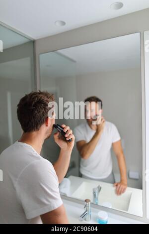 Mann Rasieren mit Elektrorasierer Trimmen seinen Bart in zu Hause Bad- Morgen Pflege Routine Menschen Konzept. Junger Mann, der den Spiegel betrachtet, der sich entwickelt Stockfoto