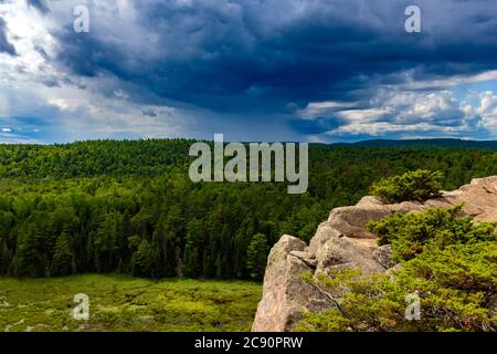 Ein wolkiger Himmel zieht sich zum Horizont hin, mit fernem Regen, der über einen Wald mit grünen Bäumen fällt. Die Wälder sind von oben mit einem felsigen cli gesehen Stockfoto