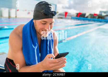 Schwimmer Mann mit Badekappe und Schutzbrille am Pool mit seinem Handy SMS auf Smartphone-App nach dem Training im Freibad. Glücklicher Athlet Stockfoto