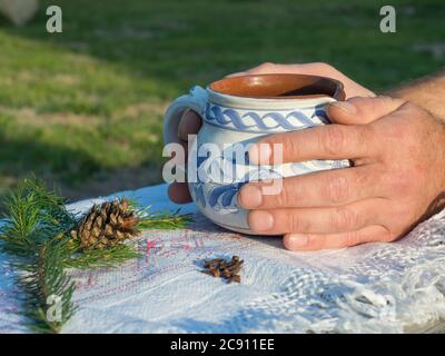 Nahaufnahme Hand des Mannes hält heißen rauchenden Tee in rustikalen weiß blau bemalten Keramik-Tasse, weiße Tischdecke, grüne Kiefer Zweige, Kegel und Nelken Gewürze Stockfoto