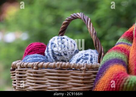 Rote, blaue und weiße Wollkugeln in einem Weidenkorb auf einer mehrfarbigen Strickdecke. Natürlicher Hintergrund. Horizontales Poster mit Platz für Kopien. Garn fo Stockfoto