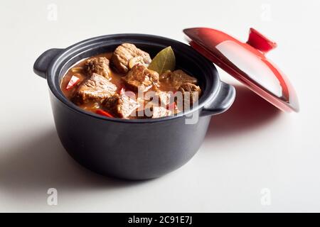 Topf mit würzigem deutschem Rindfleisch Gulasch oder Ragout serviert in einem schwarzen gusseisernen Topf mit rotem Deckel geöffnet, um den Eintopf über weiß zu offenbaren Stockfoto