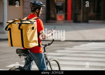 Lieferer auf dem Fahrrad. Kerl mit Rucksack und Helm hielt an der Kreuzung und schaute auf Telefon Stockfoto