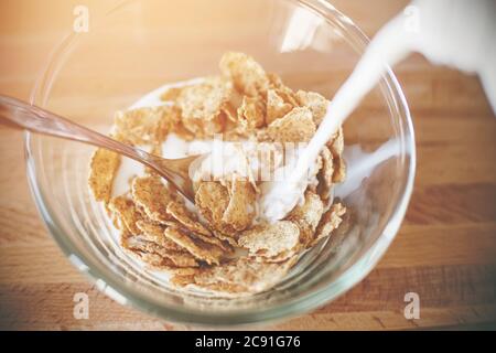 Auf dem Holztisch befindet sich eine transparente Glasschale, die Haferflocken zum Frühstück enthält, und sie werden mit frischer Kuhmilch auf einem sonnigen Mo gefüllt Stockfoto