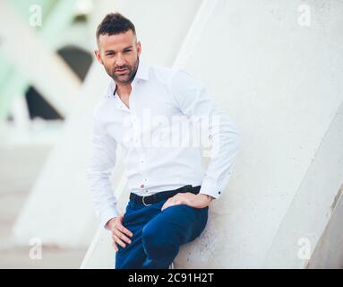Junger Mann in weißem Hemd und blauer Hose, der die Kamera anschaut, mit kurzen Haaren und kurzem Bart im Freien, der an einer weißen, schiefen Wand gelehnt ist Stockfoto