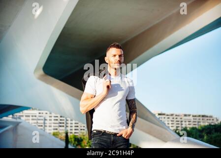 Junger Mann mit Jacke auf der Schulter und kurzen Bart zu Fuß im Freien Stockfoto