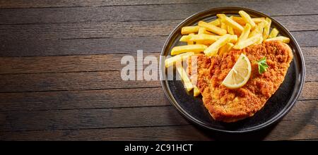 Gebratener Fisch oder Schnitzel mit Pommes auf einem Teller mit einer Zitronenscheibe auf einem Holztisch mit Copyspace in einem Panorama-Banner serviert Stockfoto