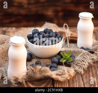 Frische Sommerbluebeere mit Blatt in der weißen Schale, zwei Joghurtflaschen auf braunem Holzhintergrund Stockfoto