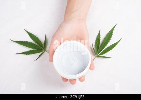 Creme in der Hand mit grünem Cannabisblatt auf weißem Hintergrund Draufsicht. Schönheit, Kosmetik für die Hautpflege mit Marihuana-Extrakt. Stockfoto