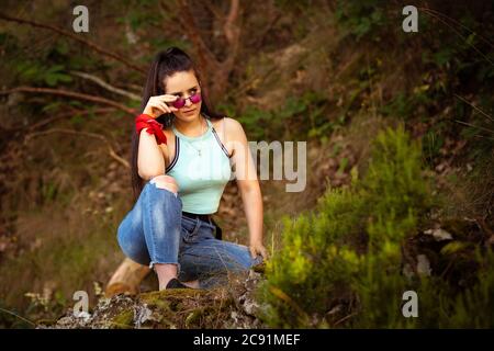 Niedriger Winkel Aufnahme einer Frau, die auf einem Felsen sitzt In einem Wald hält sie ihre Sonnenbrille an der Nase Stockfoto