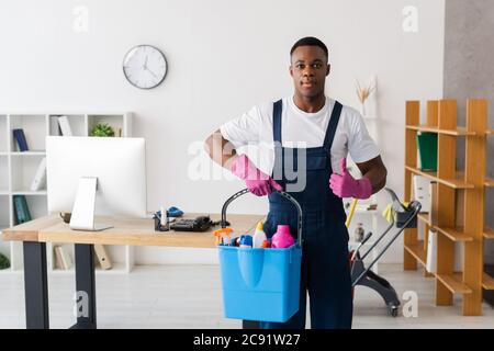 afroamerikanische Putzfrau, die Daumen hochzeigt und Eimer mit Reinigungsmitteln im Büro hält Stockfoto