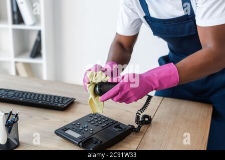 Beschnittene Ansicht des afroamerikanischen Reinigers mit Lappen beim Reinigen des Telefons auf dem Bürotisch Stockfoto