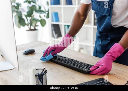 Beschnittene Ansicht der afroamerikanischen Reiniger in Gummihandschuhen Reinigung Computer-Tastatur auf Bürotisch Stockfoto