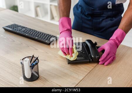 Beschnittene Ansicht der afroamerikanischen Reiniger in Arbeitskleidung und Gummihandschuhe Reinigung Telefon in der Nähe Schreibwaren und Computer-Tastatur auf dem Tisch Stockfoto