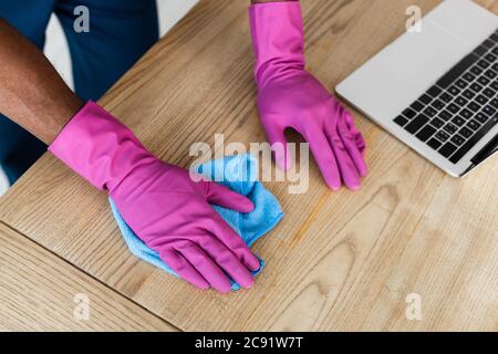 Abgeschnittene Ansicht der afroamerikanischen Reiniger in Gummihandschuhen Reinigungstisch in der Nähe Laptop im Büro Stockfoto
