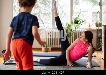 Junge Mutter trainiert Pilates, Yoga, Fitness zu Hause Blick auf den Laptop, während ihr kleiner Junge herumläuft Stockfoto