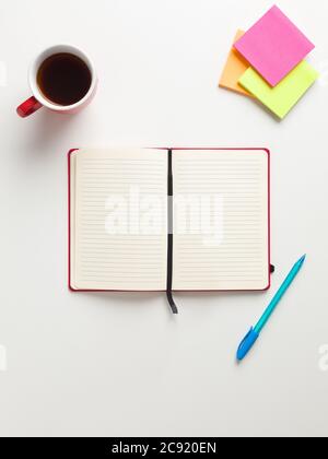 Draufsicht auf ein offenes rotes Notizbuch in der Mitte, farbige Erinnerungen in der oberen Ecke, blauer und grüner Stift und eine rote Tasse Kaffee, in weißem Backgroun Stockfoto