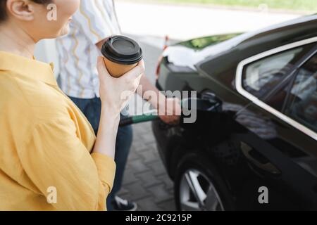 Beschnittene Ansicht einer Frau, die Kaffee hält, um zu gehen, während der Mann an der Tankstelle Auto anschaufelt Stockfoto