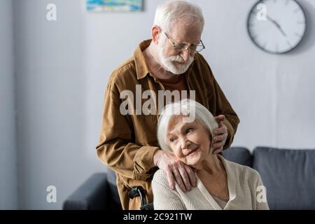 Lächelnde ältere Frau im Rollstuhl sitzend, während der Ehemann die Hand auf der Schulter hielt und die Haare berührte Stockfoto