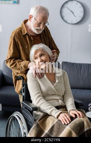 Lächelnde, verträumte ältere Frau, die im Rollstuhl sitzt, während der Mann die Hand auf der Schulter hält und Haare berührt Stockfoto