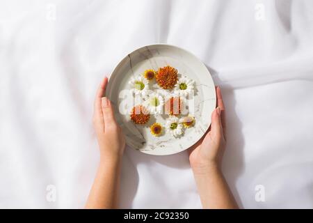 Draufsicht auf Voile Stoff Hintergrund mit weiblichen Händen halten einen Teller mit schwebenden Blumen Stockfoto