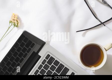 Draufsicht auf Morgentee im Voile Stoff Hintergrund mit einem Laptop, Tasse Tee, Gläser und Blumen mit Platz für Text Stockfoto
