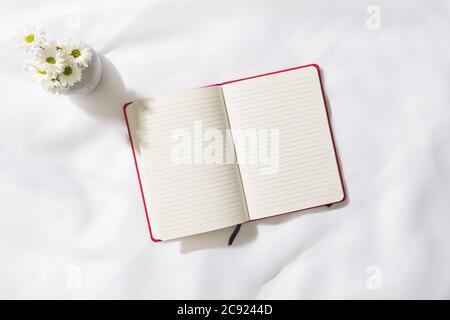 Draufsicht der Morgenszene im Voile-Stoff-Hintergrund mit einem roten Notizbuch in der Mitte und einer Vase mit weißen Blumen, mit Platz für Text Stockfoto