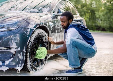 Luxus elektrische moderne Auto in Seifenschaum im Freien bei Waschservice. Seitenansicht des hübschen jungen afrikanischen Mann mit grünen Mikrofaser Auto Waschhandschuh für Stockfoto