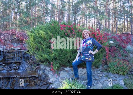 Reife Frau in Jeans und eine bunte Bluse vor einem Hintergrund aus grünen Sträuchern, roten Blättern und einem kleinen improvisierten Wasserfall Stockfoto