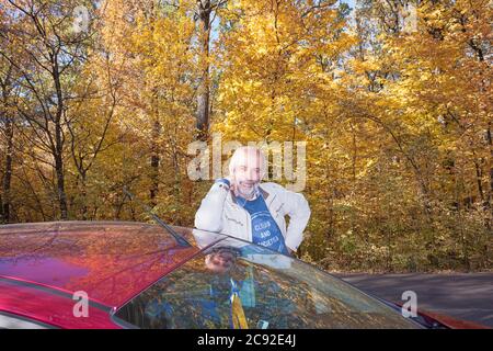 Ein Mann von reifem Alter ruht auf dem Dach eines Autos, das auf der Seite einer Waldstraße geparkt ist. Hintergrund - schöne gelbe Laub von Bäumen Stockfoto