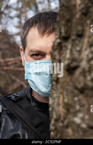 Ein Mann in einer medizinischen Maske steht hinter einem Baum. Der Kampf gegen das Coronavirus. Stockfoto