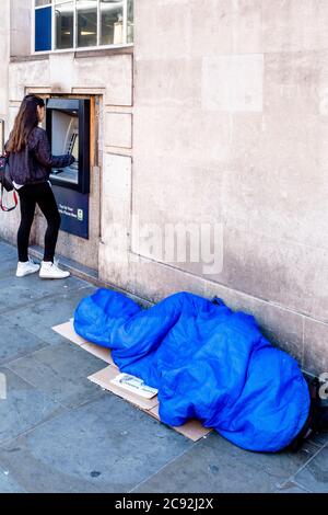 Eine obdachlose Person schläft in EINEM Schlafsack, während EINE junge Frau Geld an einem Geldautomaten in der Borough High Street, London, England abzieht. Stockfoto