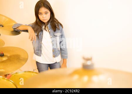 Ein Porträt eines niedlichen asiatischen Grundschulmädchen mit langen Haaren und einer Denim-Jacke, die mit einer Trommel in einem Klassenzimmer mit einer weißen Wand steht. Stockfoto