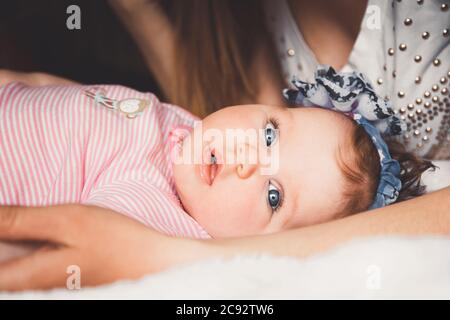 Nettes 3 Monate altes Baby Mädchen, das sich auf einem Bett in den Händen der Mutter legt. Mutter und Kind auf einem weißen Bett zu Hause. Große offene Augen. Stockfoto