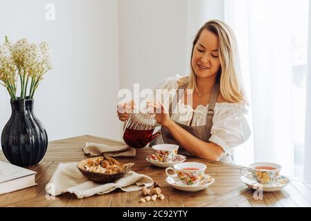 Blonde Frau gießt Hibiskustee in Tassen, während sie sitzt Tabelle Stockfoto