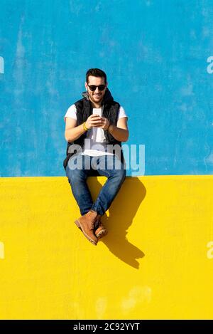 Junger bärtiger Mann mit Sonnenbrille sitzt an der gelben Wand und benutzt Telefon Stockfoto